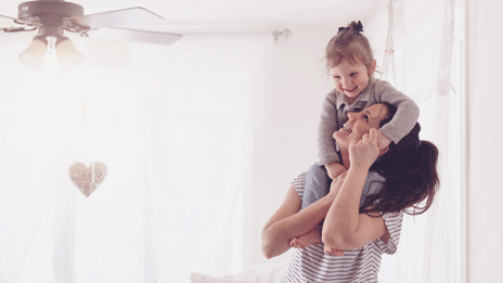 Young mother with her toddler daughter on her shoulders in their bright living room.