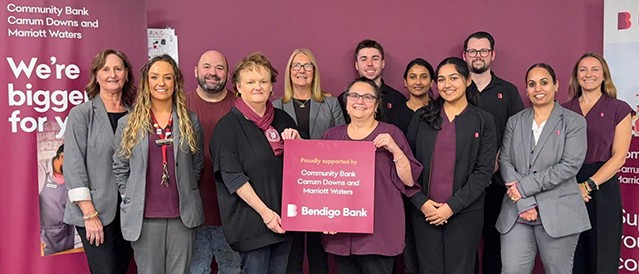 A group of 12 branch staff member standing in front of a plum-coloured wall holding a Bendigo Bank sign.