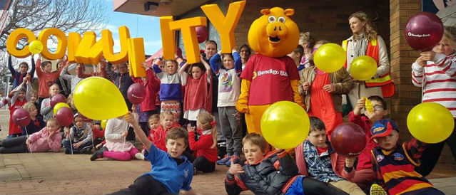 Children with burgundy and gold balloons and big yellow letters spelling the word 'community' sitting and standing outside Edenhope Community Bank Branch.
