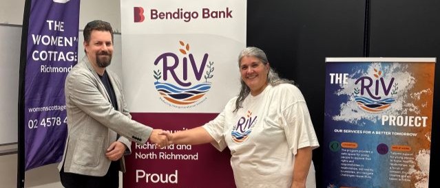 A man and a woman shaking hands in front of Bendigo Bank and RIV Project signs.