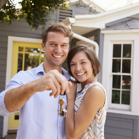 Stock image of a couple standing out the front of a home holding keys representing first home buyers.