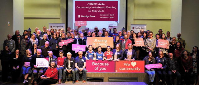 A group of local people at a community grant event holding signs with different messages including sharing is caring, because of you, we love community, a shared return, thanks to you and better together.  