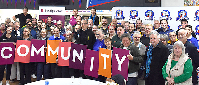A large group of people standing in from of a Bendigo Bank banner, holding placards that spell 'community'