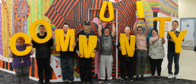 Araluen members standing holding yellow COMMUNITY letters infront of painted mural at the Diamond Creek Disability Support Centre.