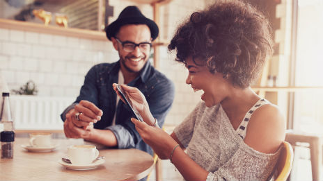 Man and a woman sitting at a table, the woman is looking at her phone with a huge smile and the man is looking at the woman laughing