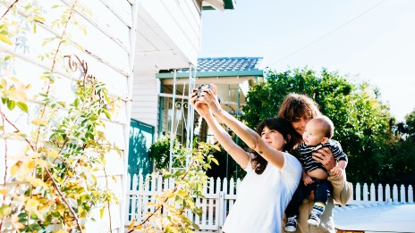 mother and father standing together in front of a white weatherboard house smiling cuddling their baby