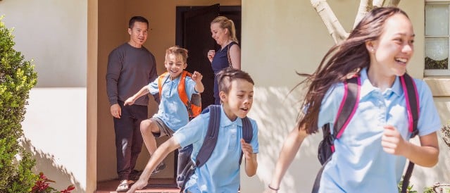 Three excited school children running outside their front door with backpacks on.