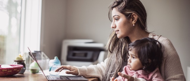 A person sitting down at a table using their laptop while holding a baby.