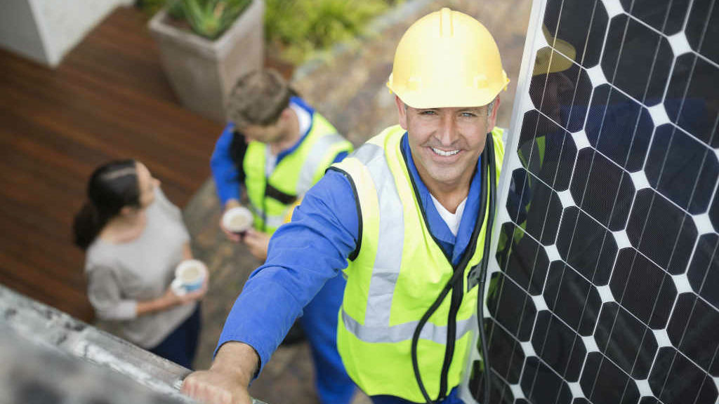 A tradesman in high vis work vest and hard hat on ladder holding solar panel.